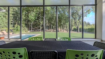 Une pièce dotée d’une grande fenêtre donnant sur une piscine et un espace verdoyant avec des arbres. On y trouve une table recouverte d’une nappe noire et des chaises vertes.