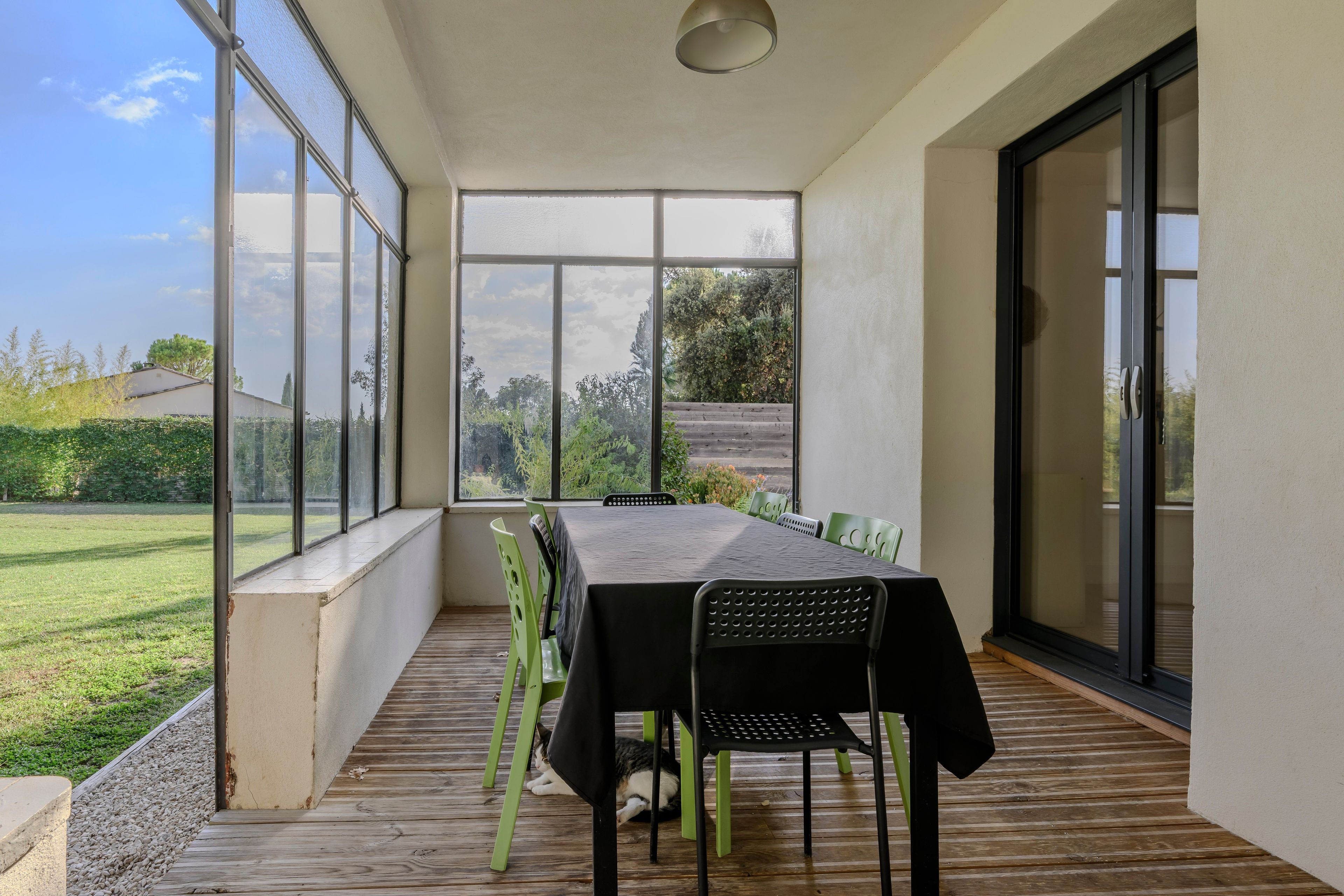 Varanda coberta com mesa e cadeiras, grandes janelas e vista para um jardim.