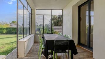 Varanda coberta com mesa e cadeiras, grandes janelas e vista para um jardim.