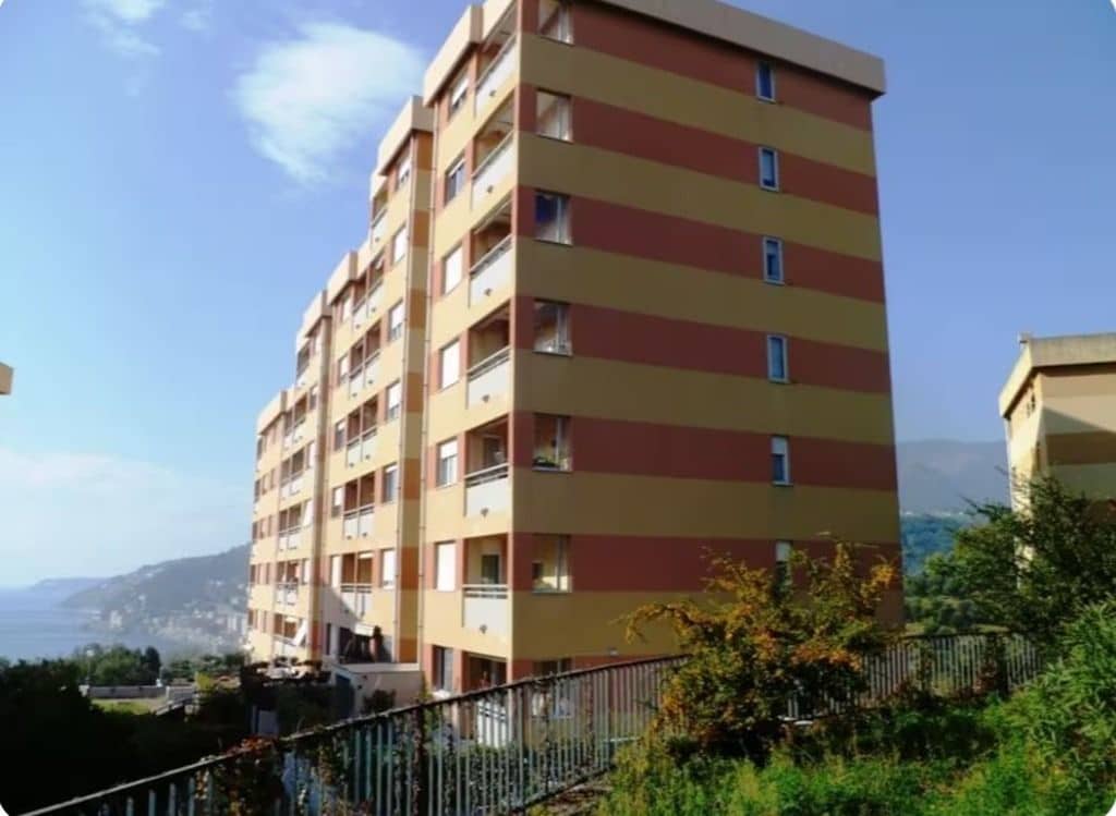 A multi-story residential building with balconies, located in a hilly area with a clear sky.