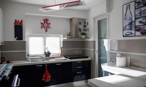 A modern kitchen with a central island, a window with blinds, and a red decorative wall piece. - A Window on the Sea | House with a Sea View on the Genoa Riviera (Genova)