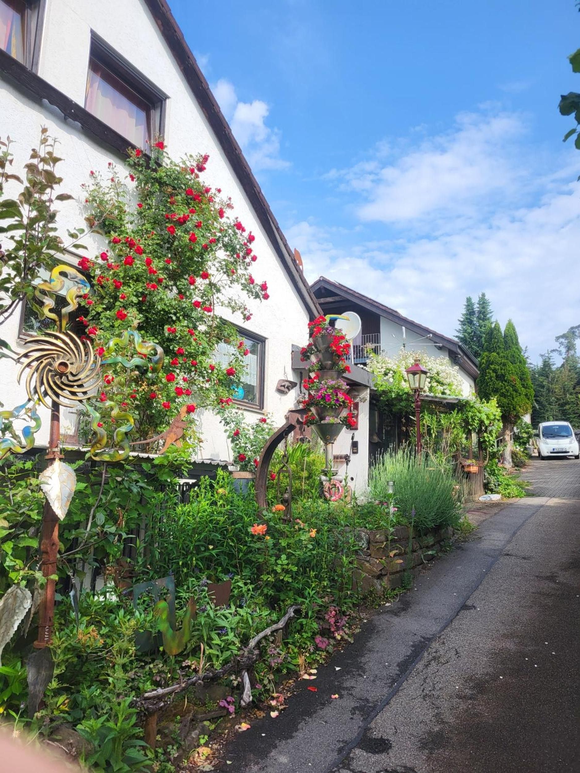 A residential street with a white house featuring a garden with red flowers and a decorative metal sculpture.