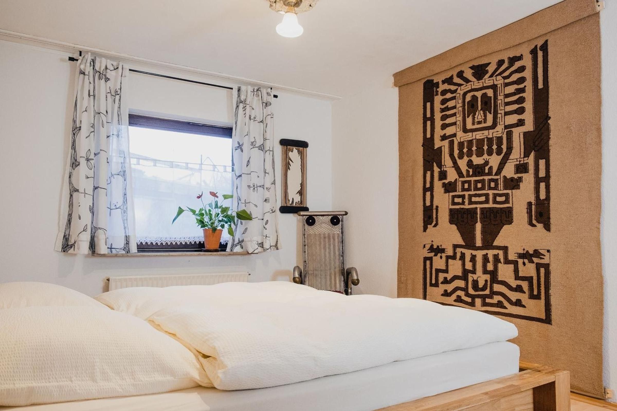 Ein Schlafzimmer mit Bett, einem Fenster mit Vorhängen, einem Heizkörper, einer Topfpflanze und Wandteppich.