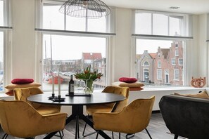 Een moderne kamer met een ronde tafel, gele stoelen en een zwarte bank. Er zijn grote ramen met uitzicht op gebouwen en een stadsbeeld.