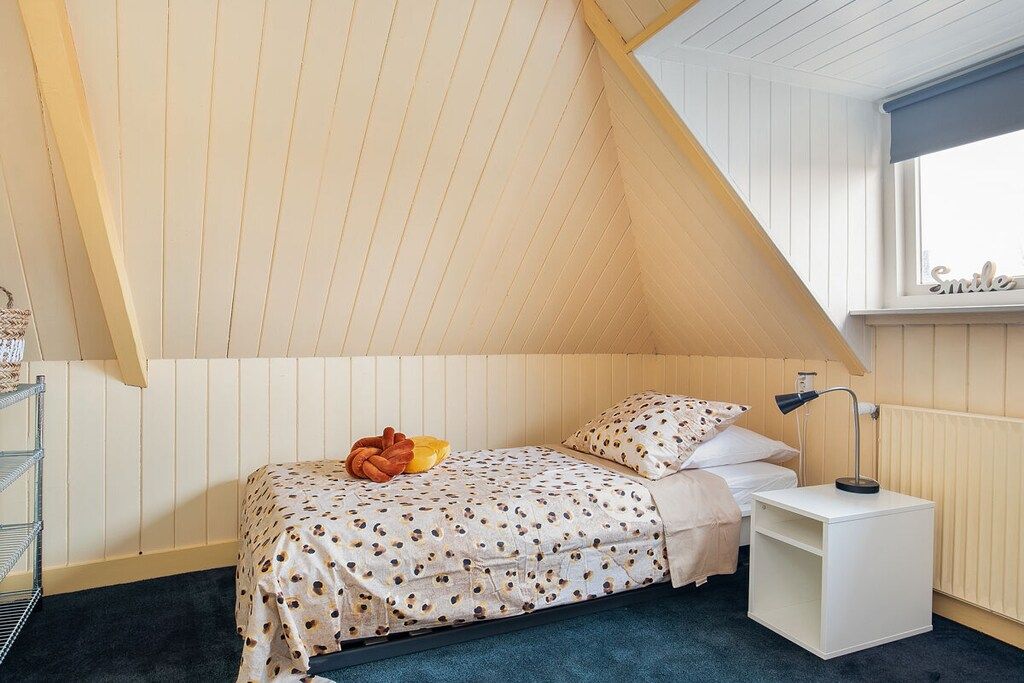 Een slaapkamer met een schuine kap, een bed met een stippenbedekking, een bedlampje en een raam met een 'Smile'-bordje.