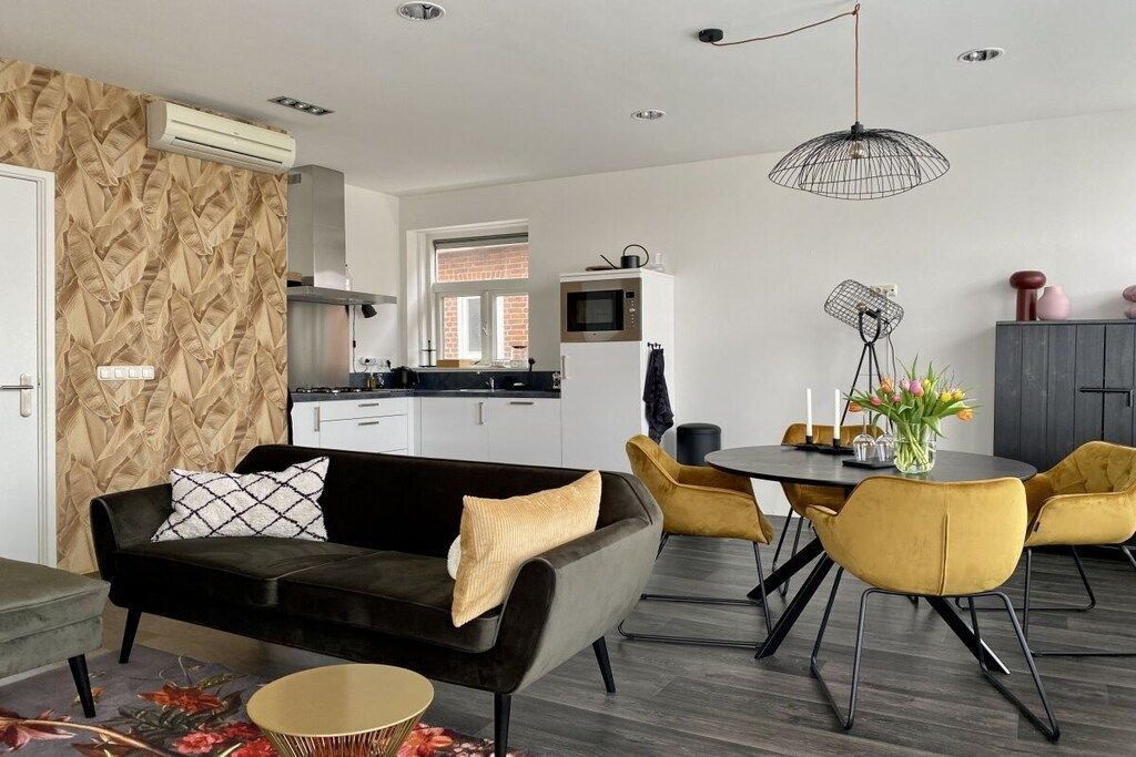 Een moderne woonkamer met een zwarte bank, gele stoelen, een eettafel en een keuken op de achtergrond.