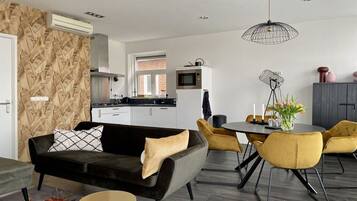 Een moderne woonkamer met een zwarte bank, gele stoelen, een eettafel en een keuken op de achtergrond.