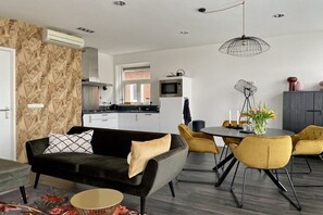 Een moderne woonkamer met een zwarte bank, gele stoelen, een eettafel en een keuken op de achtergrond.