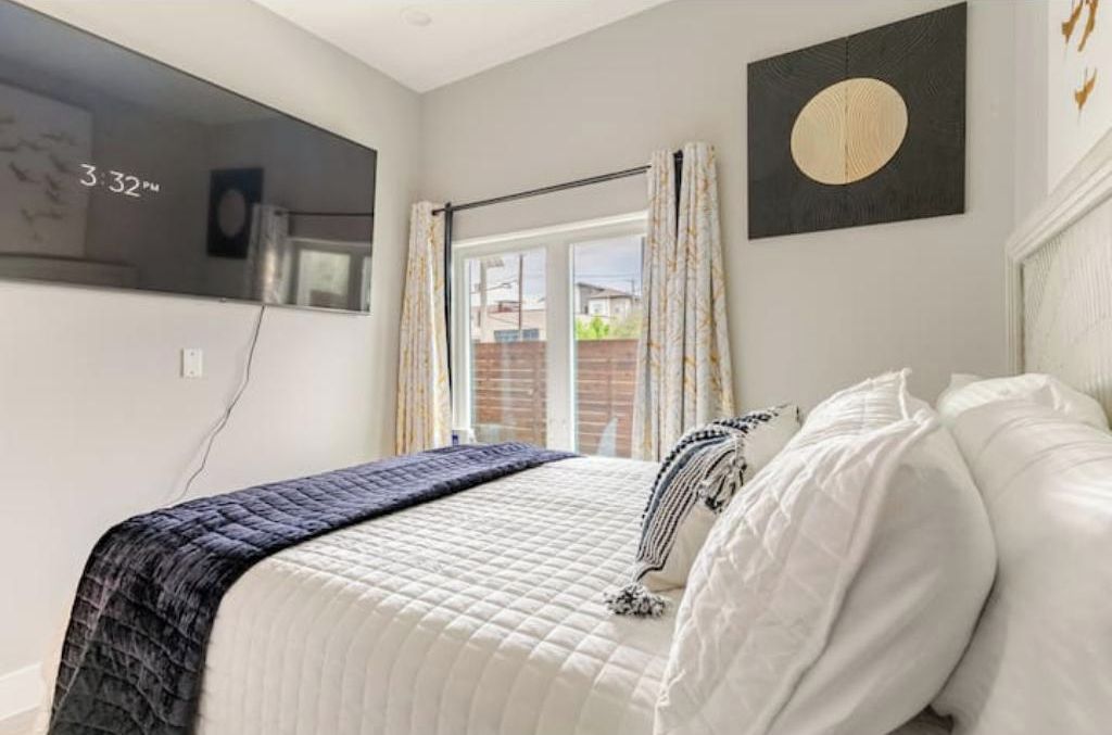 A bedroom with a bed, a window with curtains, a wall clock, and a framed artwork.