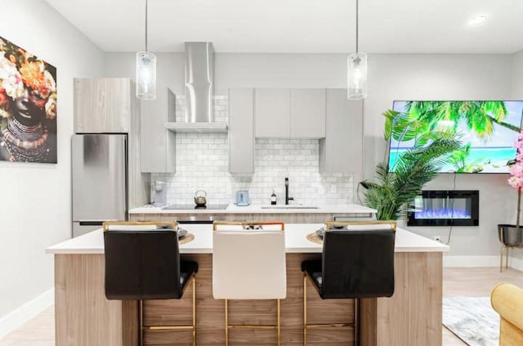 A modern kitchen with a central island, two bar stools, and a flat-screen TV mounted on the wall.