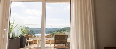 Habitación de hotel con un amplio ventanal corredero que da a un balcón exterior con vistas a un lago y colinas.