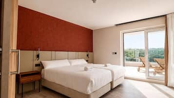A hotel room with a bed, a nightstand, a wooden bench, a red brick accent wall, and a sliding glass door leading to a balcony with outdoor furniture.