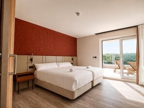 A hotel room with a bed, a nightstand, a wooden bench, a red brick accent wall, and a sliding glass door leading to a balcony with outdoor furniture.