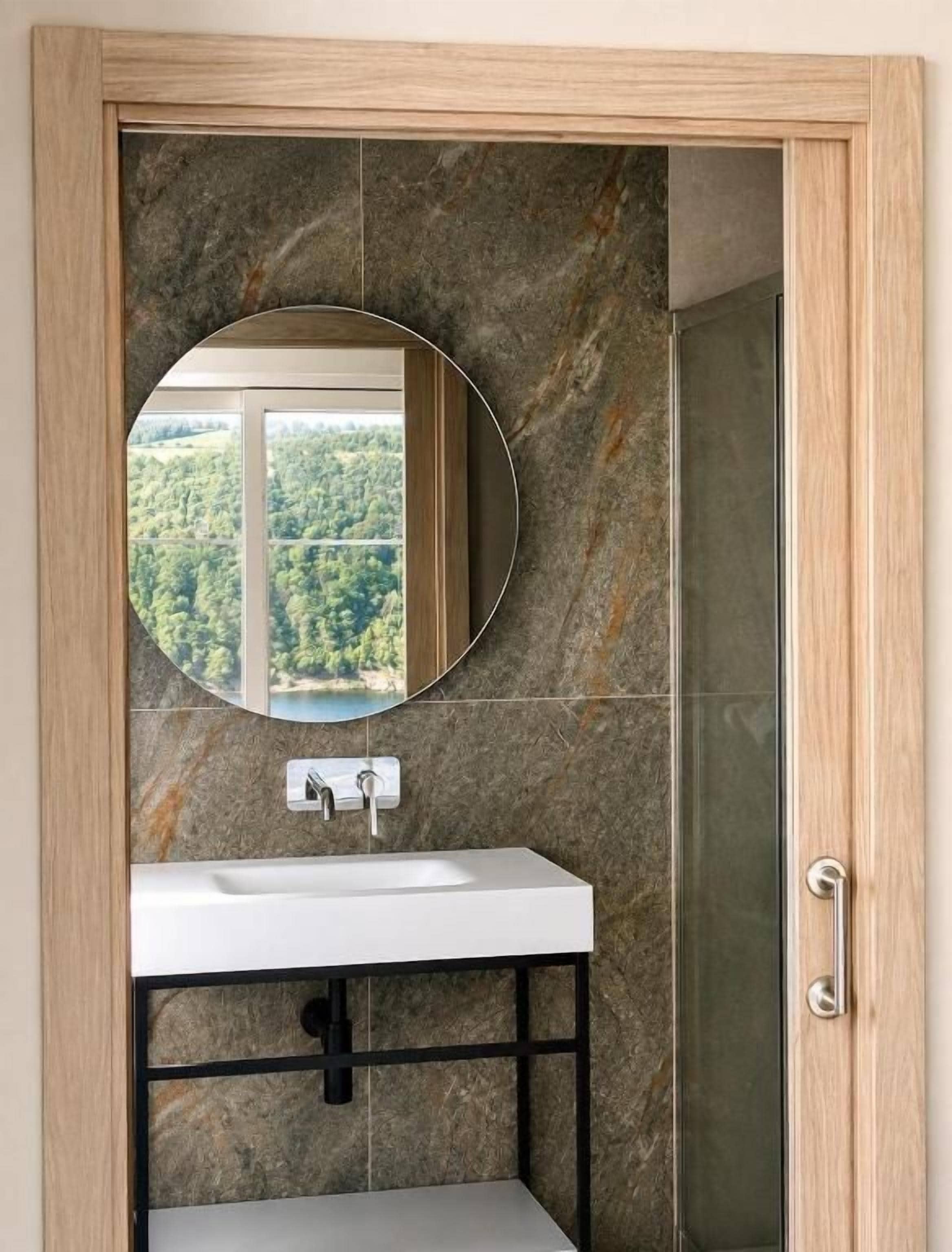 Une salle de bain avec un lavabo blanc, un miroir rond et une vue sur une forêt par la fenêtre.