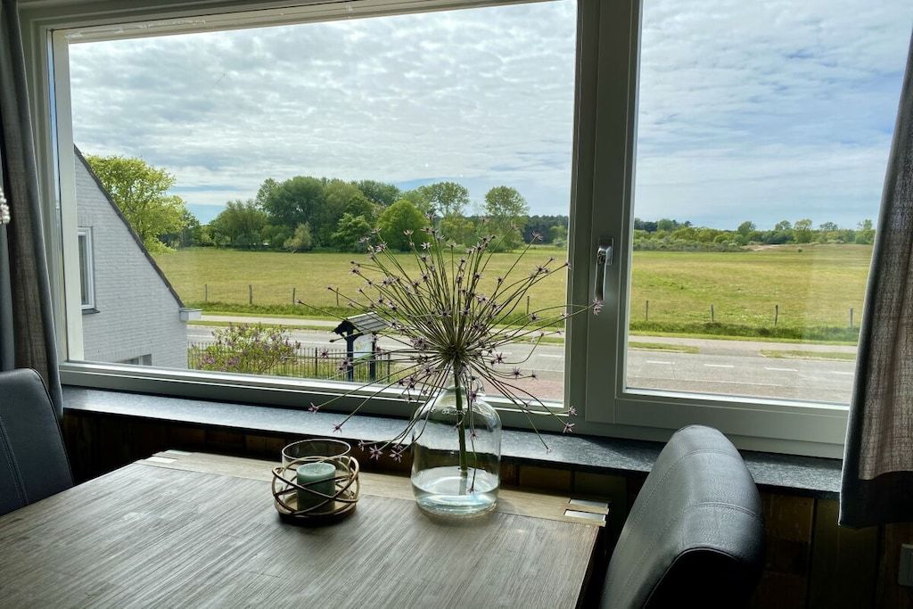 A room with a view of a green field and trees, a window with a vase of flowers, and a table with a candle and a coaster.