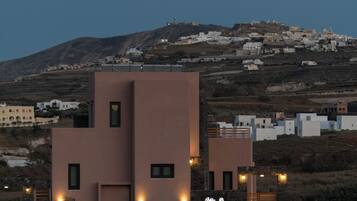 A modern building with a flat roof and multiple windows, surrounded by a hilly landscape with other buildings.