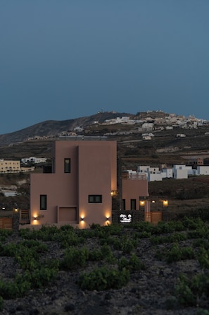 A modern building with a flat roof and multiple windows, surrounded by a hilly landscape with other buildings.
