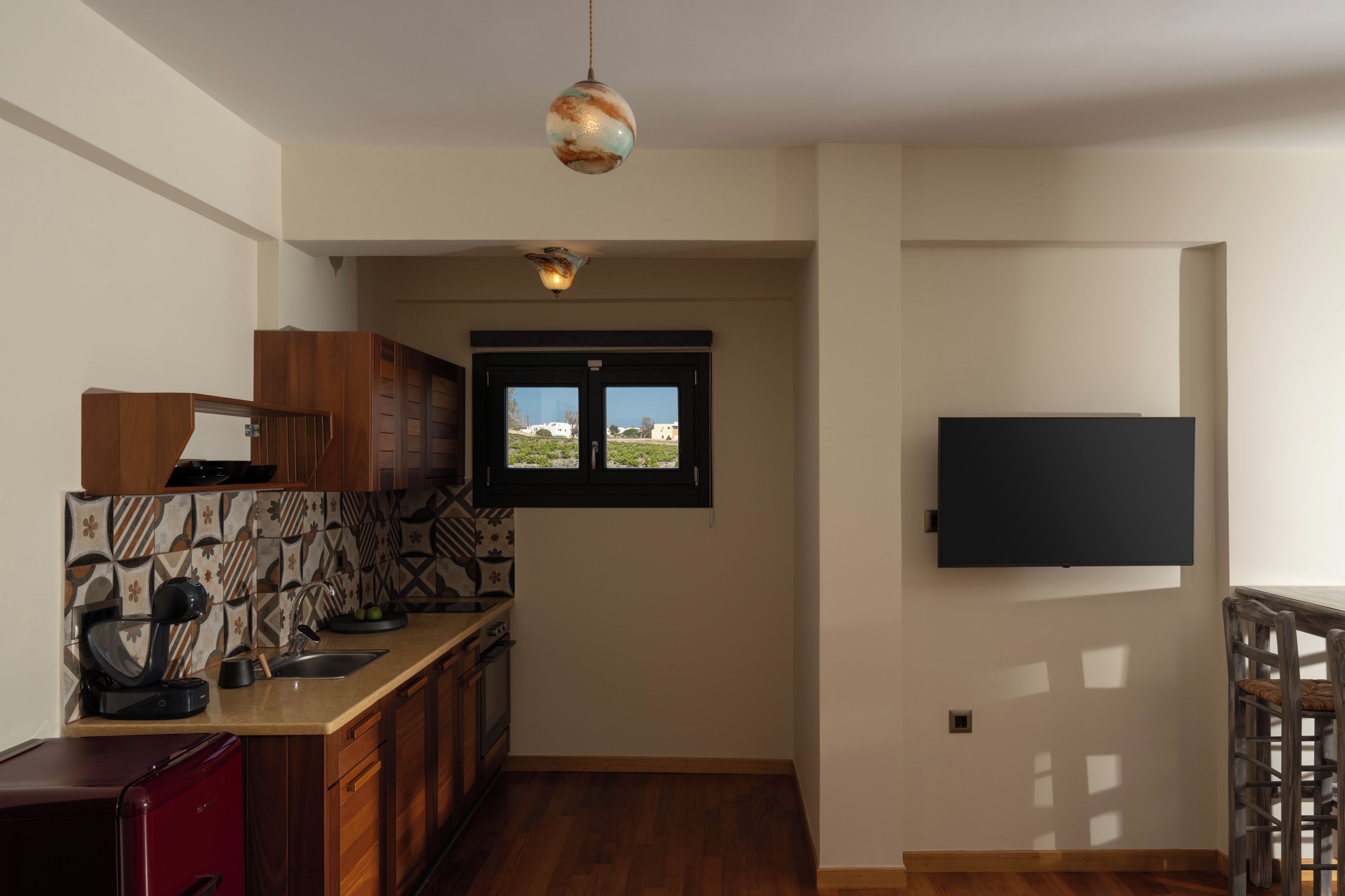 Een compacte keuken met een inbouwoven, een magnetron en een spoelbak. Er is een raam met uitzicht naar buiten en een flatscreen televisie bevestigd aan de muur.