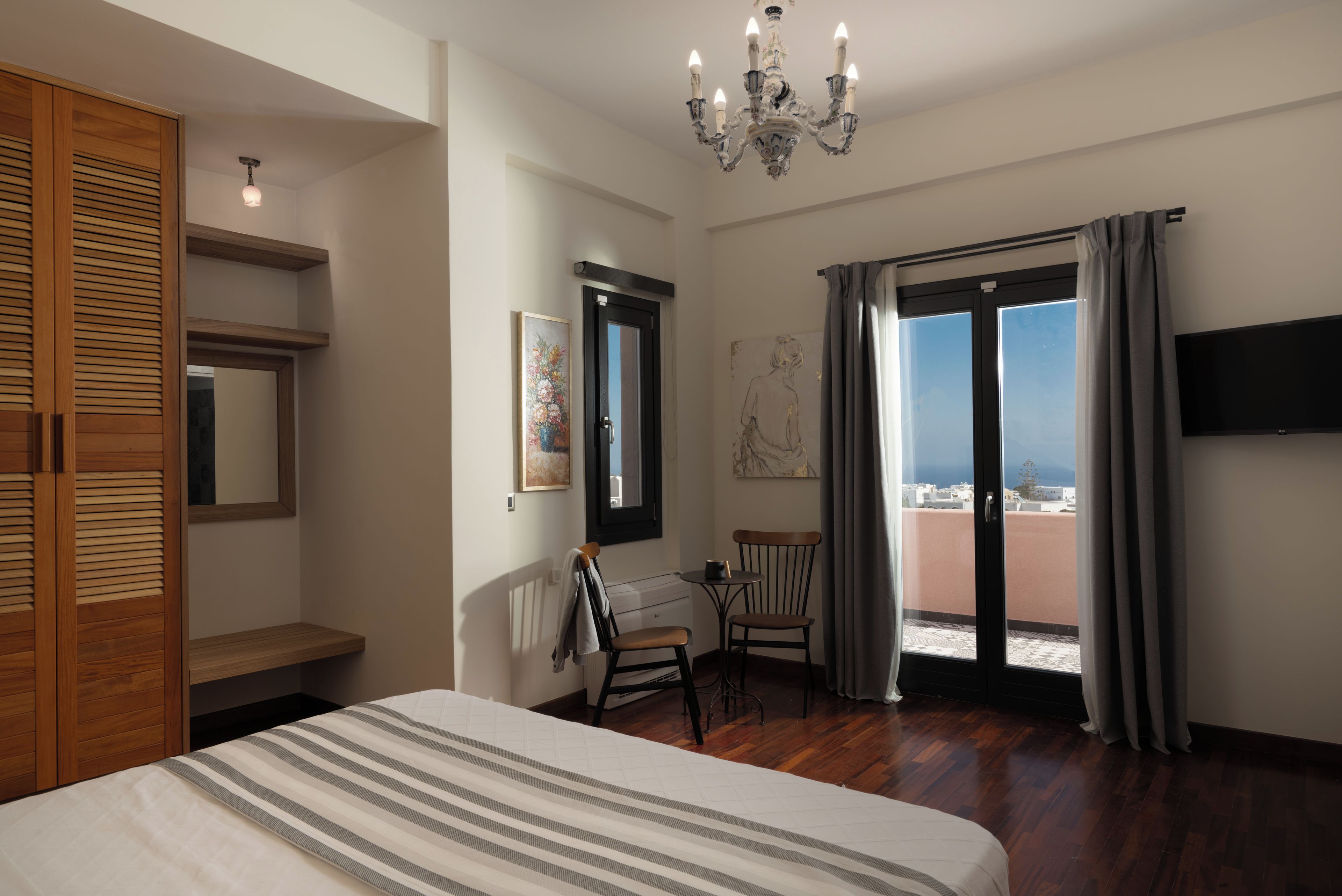 Ein Hotelzimmer mit Bett, einem Holzschrank, einem Schreibtisch, einem Sessel, einem Balkon mit Blick und einer Kristallleuchter.