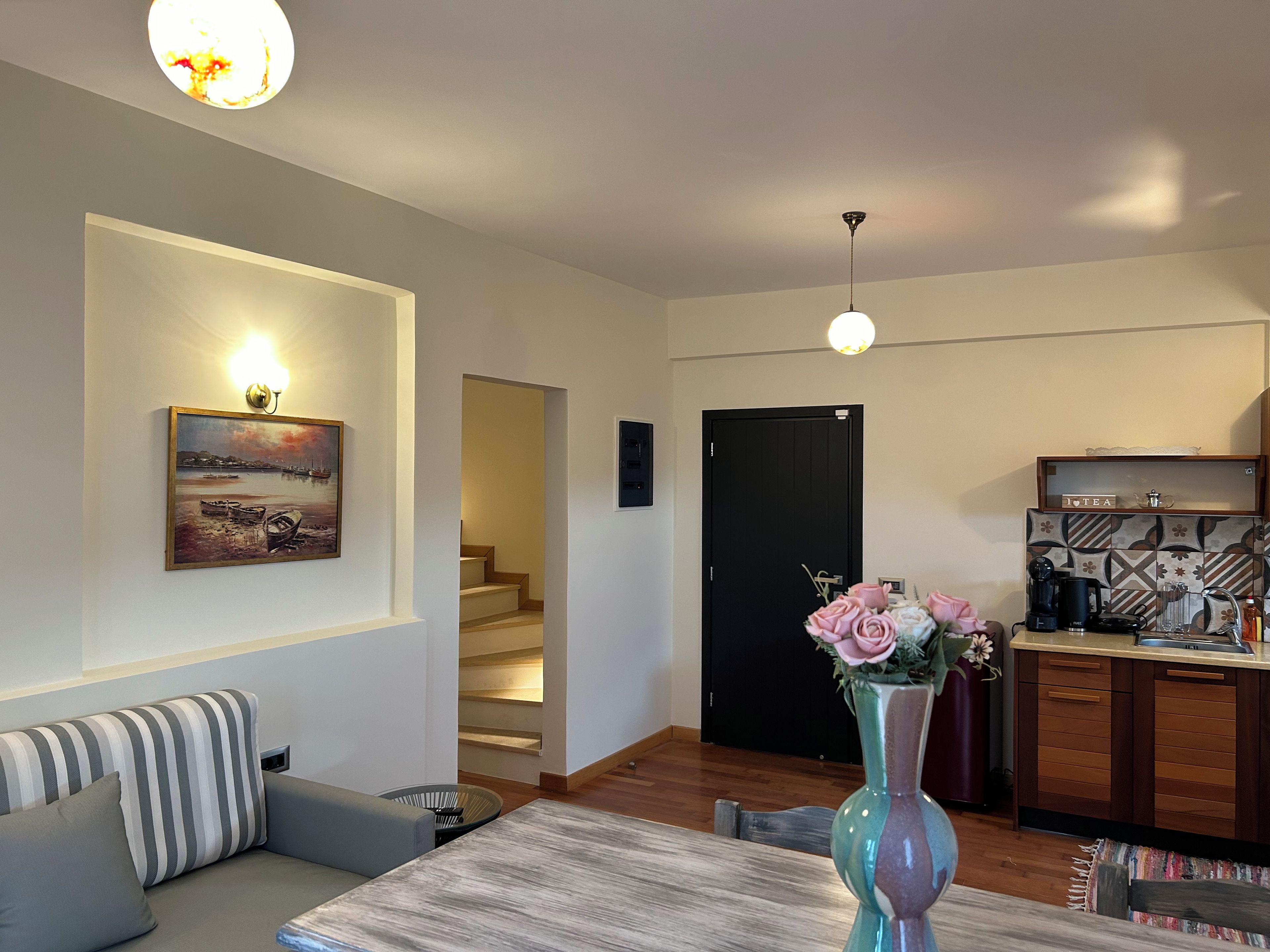 Una sala moderna con un sofá, una mesa de comedor, un jarrón con flores, una zona de cocina con cafetera y una escalera que lleva a un piso superior.