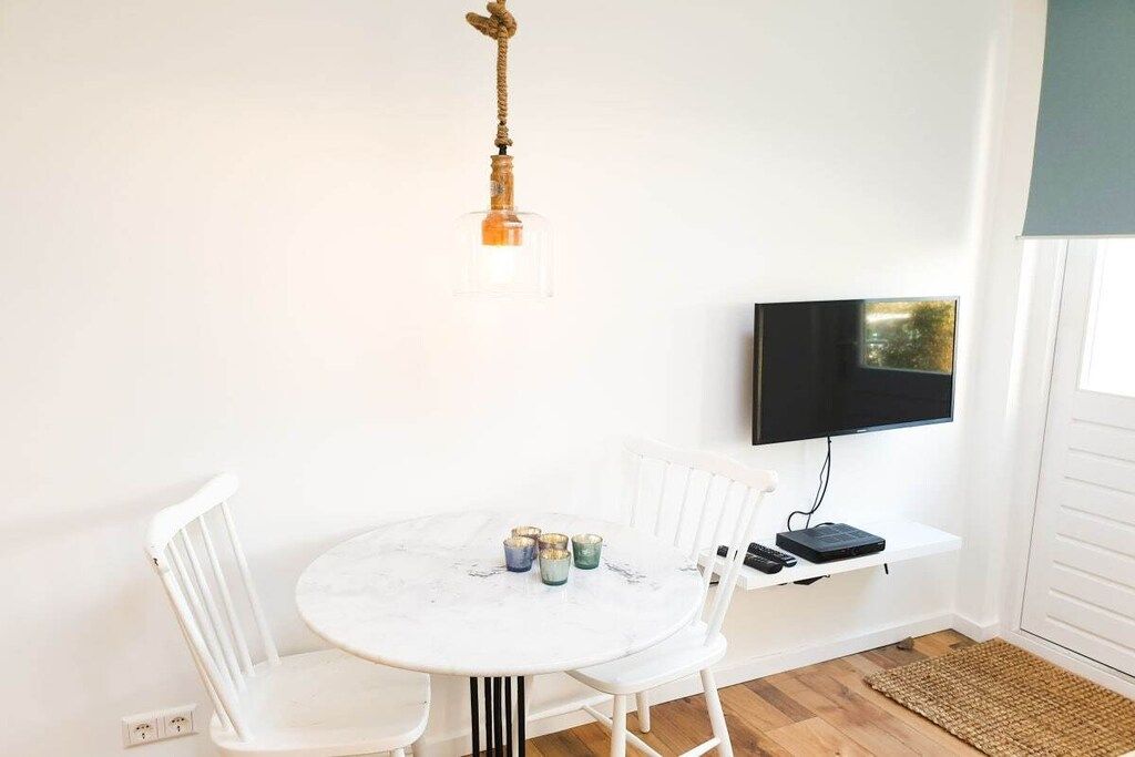 Ein Zimmer mit einem runden Tisch, weißen Stühlen, einem an der Wand befestigten Fernseher und einem kleinen Regal mit einer Fernbedienung.