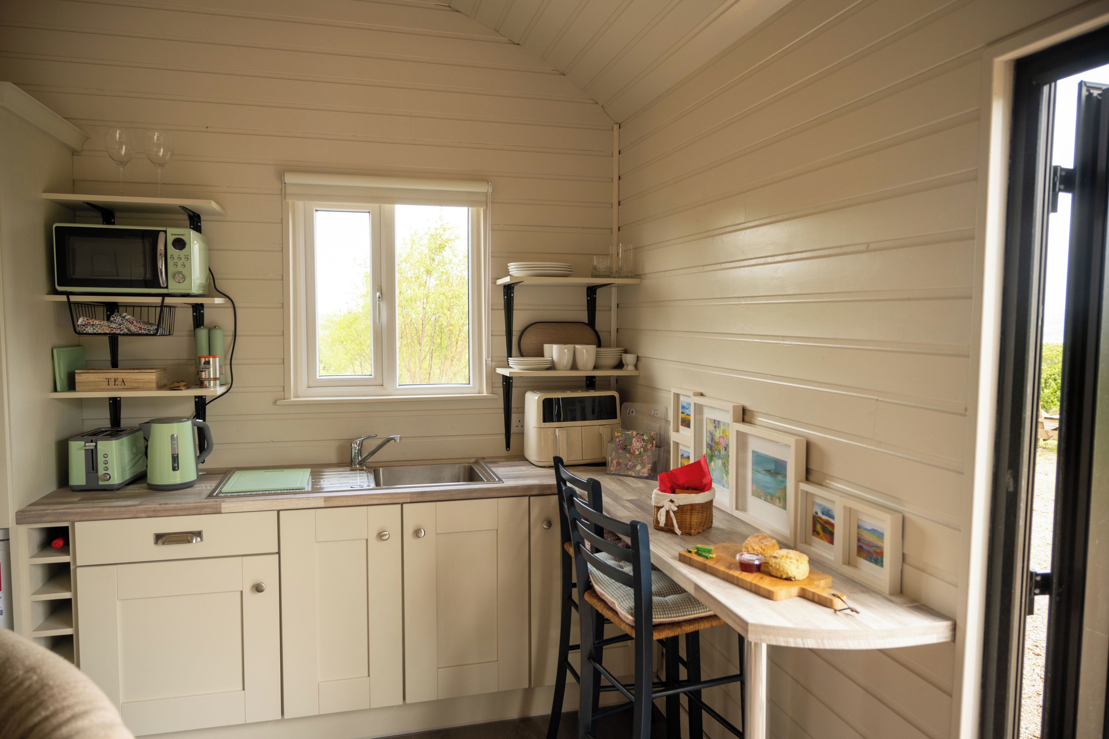 A compact kitchen with a sink, a microwave, and a toaster. There is a window providing natural light, and a dining area with a chair and a table.