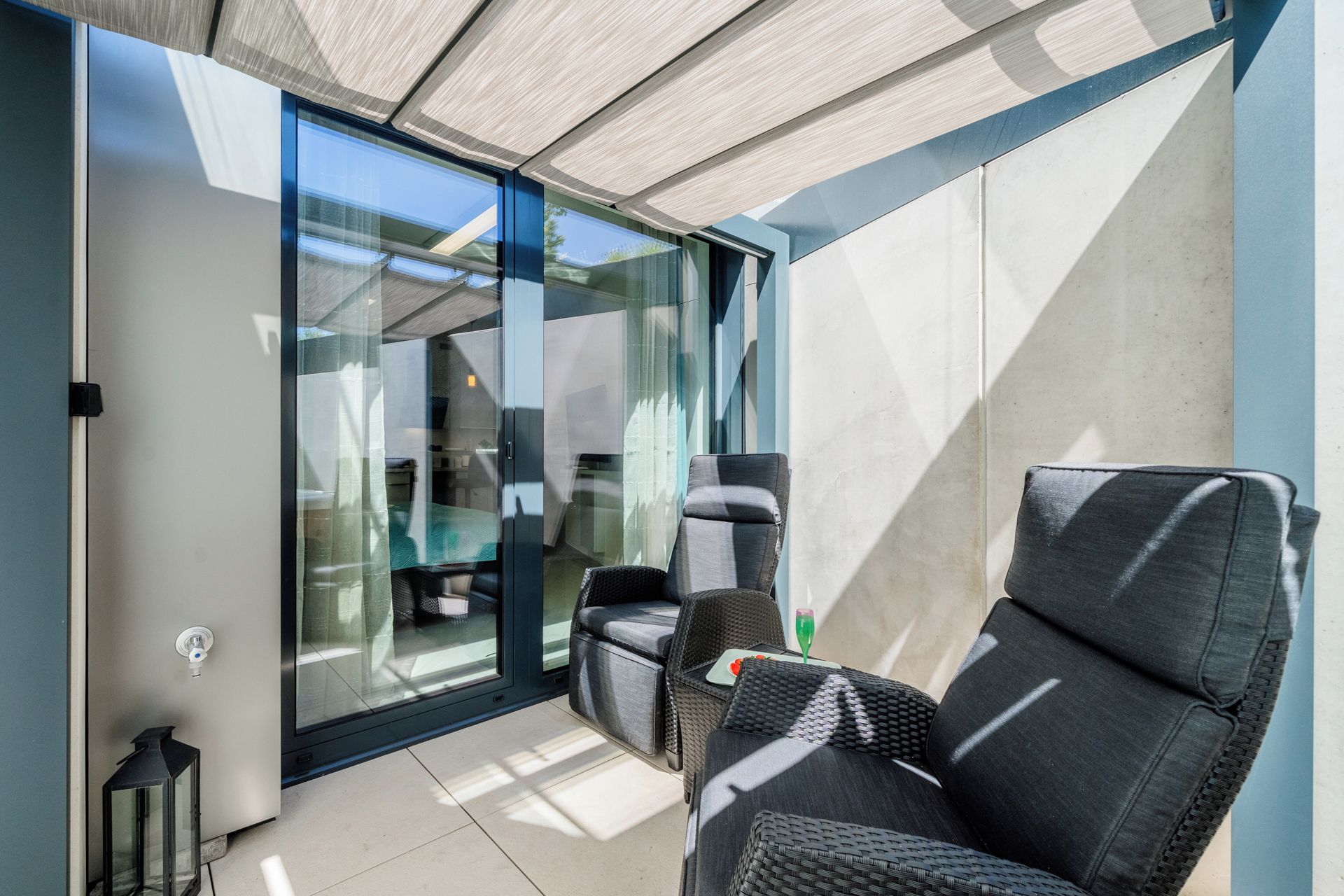 Ein moderner Außenbereich mit einer Glastür, die auf eine Terrasse führt, ein Korbstuhl und ein kleiner Tisch mit einem Glas.