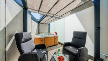 A sunlit outdoor seating area with two chairs, a small table with a bowl of strawberries, and a hot tub.