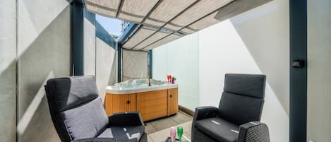 A sunlit outdoor seating area with two chairs, a small table with a bowl of strawberries, and a hot tub.