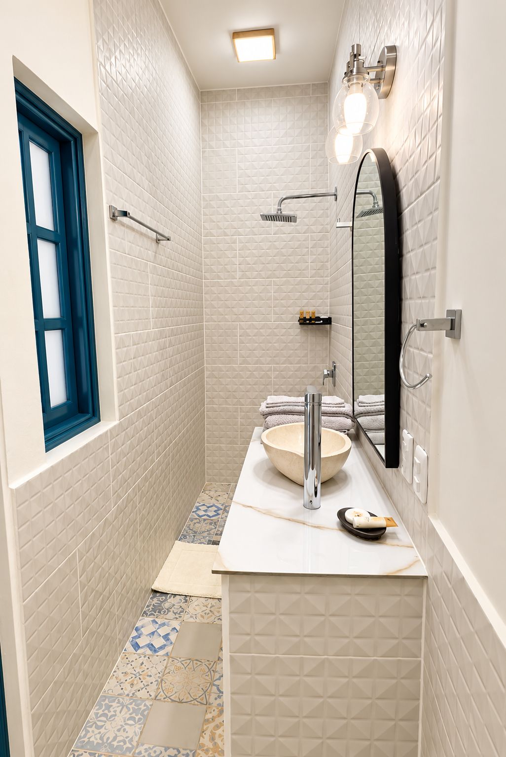 Une salle de bain avec un mur recouvert de carreaux blancs, un grand miroir, un lavabo et un espace douche.