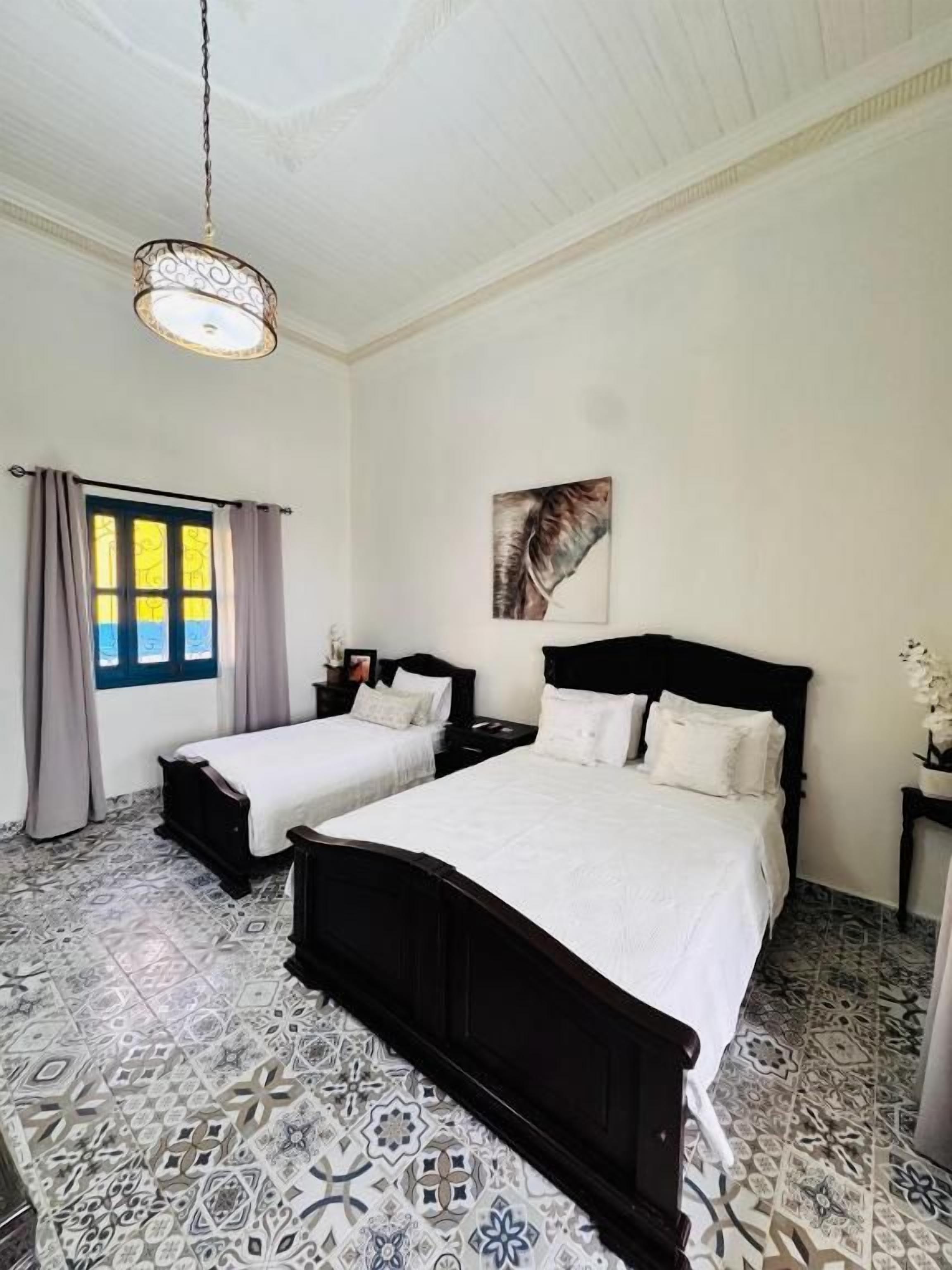 Une chambre avec deux lits, un lustre, une fenêtre avec des rideaux et des carreaux de sol à motifs.