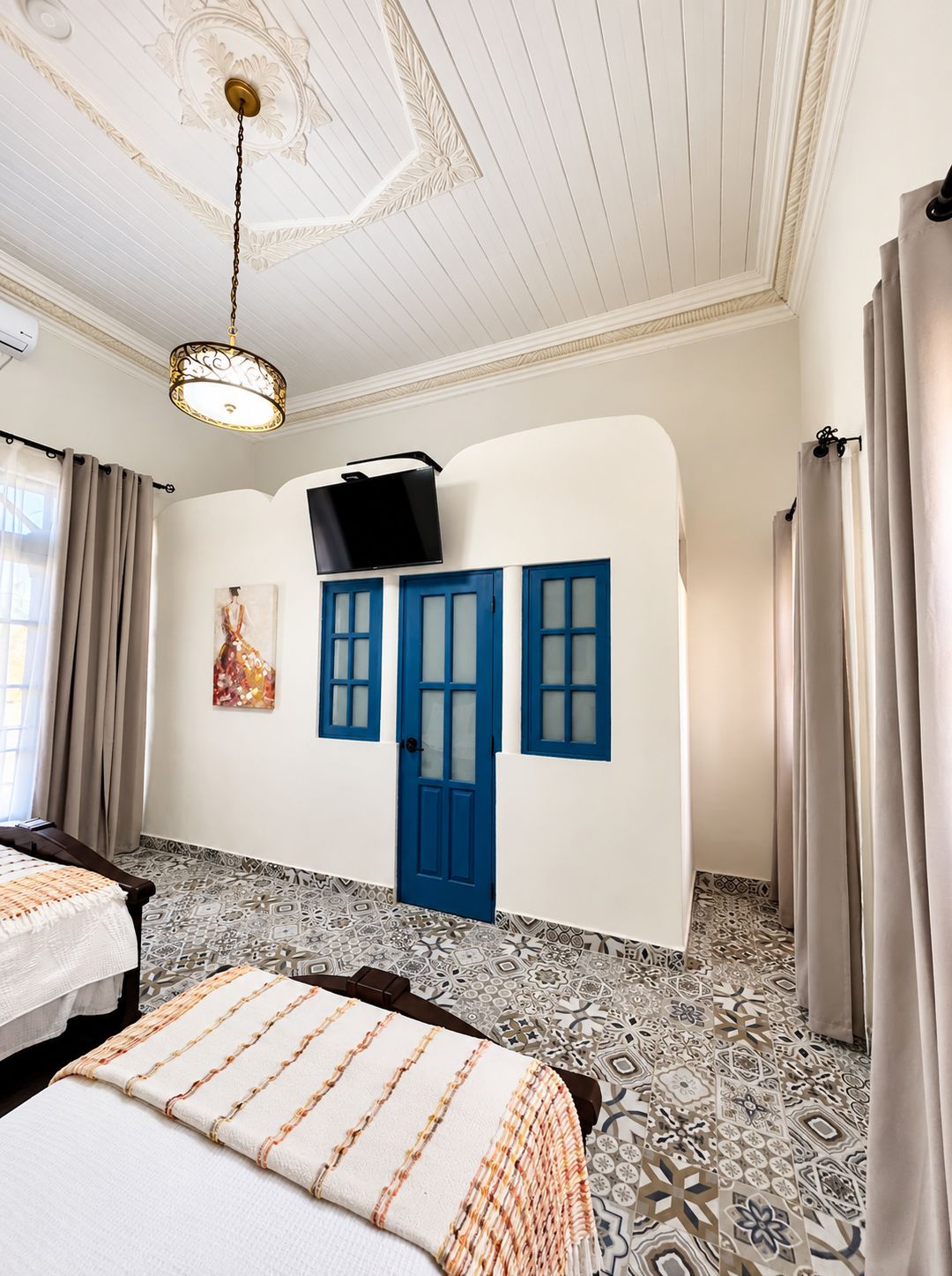 Quarto de hotel com cama, televisão presa na parede, porta azul, ladrilhos de chão estampados e teto com design decorativo.