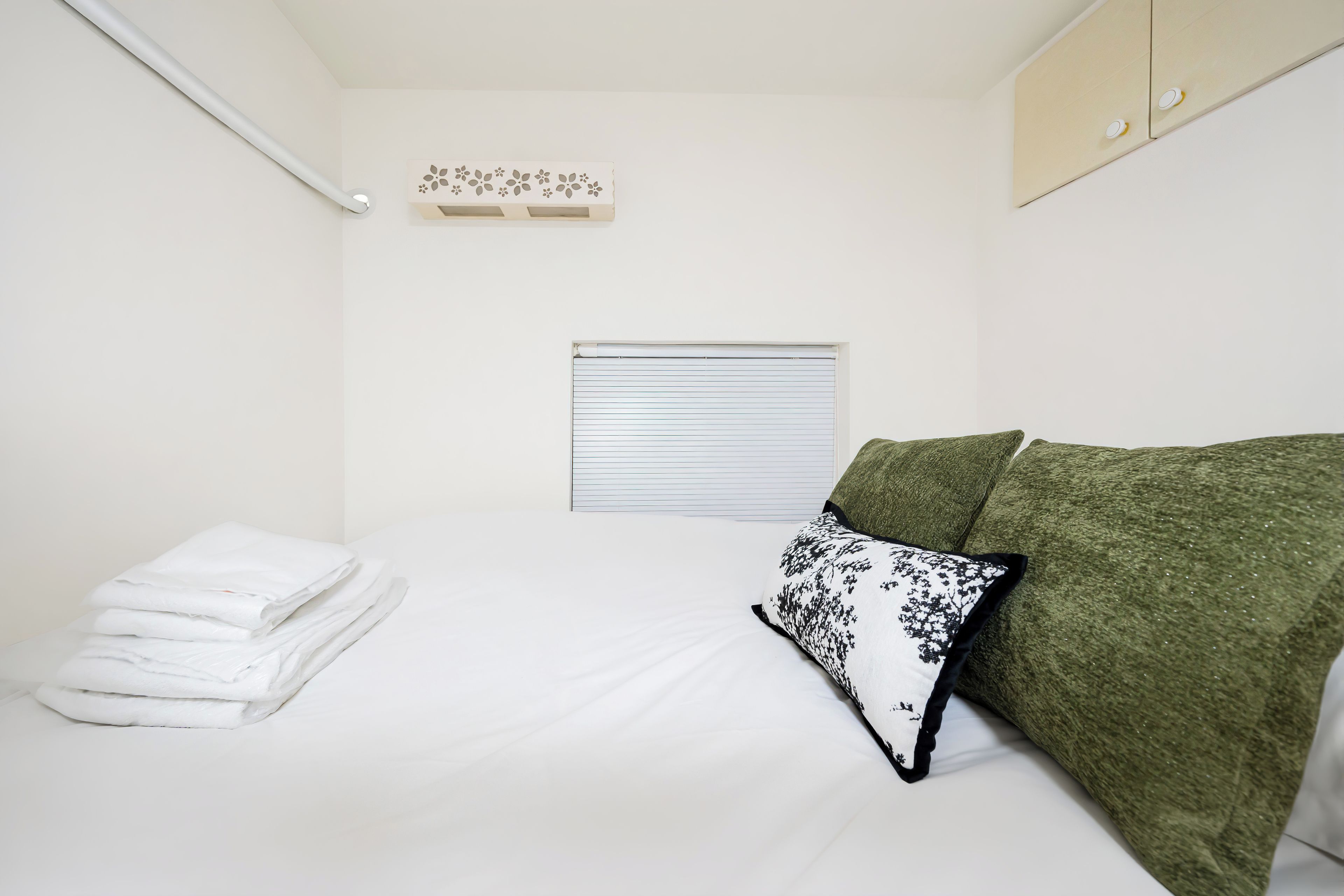 Un dormitorio pequeño y ordenado con una cama, una pila de toallas blancas, un ventanal con persianas cerradas y un sofá verde con un cojín estampado.