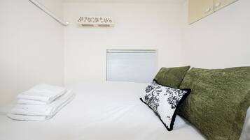 Un dormitorio pequeño y ordenado con una cama, una pila de toallas blancas, un ventanal con persianas cerradas y un sofá verde con un cojín estampado.