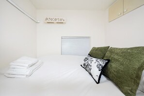 A small, neatly arranged bedroom with a bed, a stack of white towels, a window with closed blinds, and a green couch with a patterned pillow.
