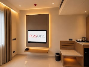 Una habitación de hotel moderna con una televisión de pantalla plana montada en la pared, una pequeña zona de estar, una kitchenette y un cesto de basura.