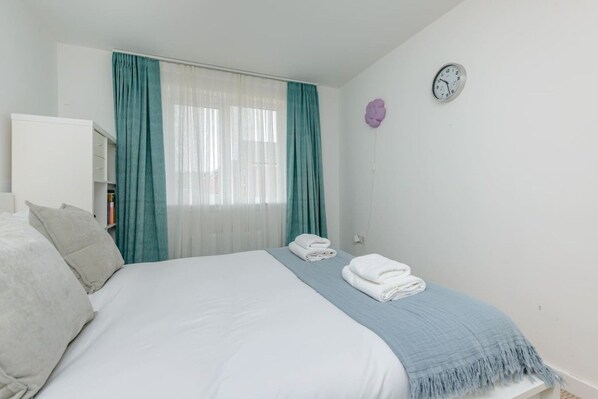 A bedroom with a bed, a bedside table with towels, a clock, and a window with curtains. - Stylish 5-Bed, 2-Bath House Near Edinburgh Centre (Edinburgh)