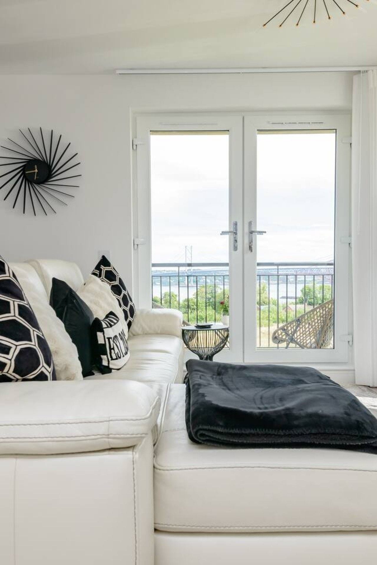 A white sofa with patterned pillows, a black throw blanket, and a view of a balcony with a railing and greenery.