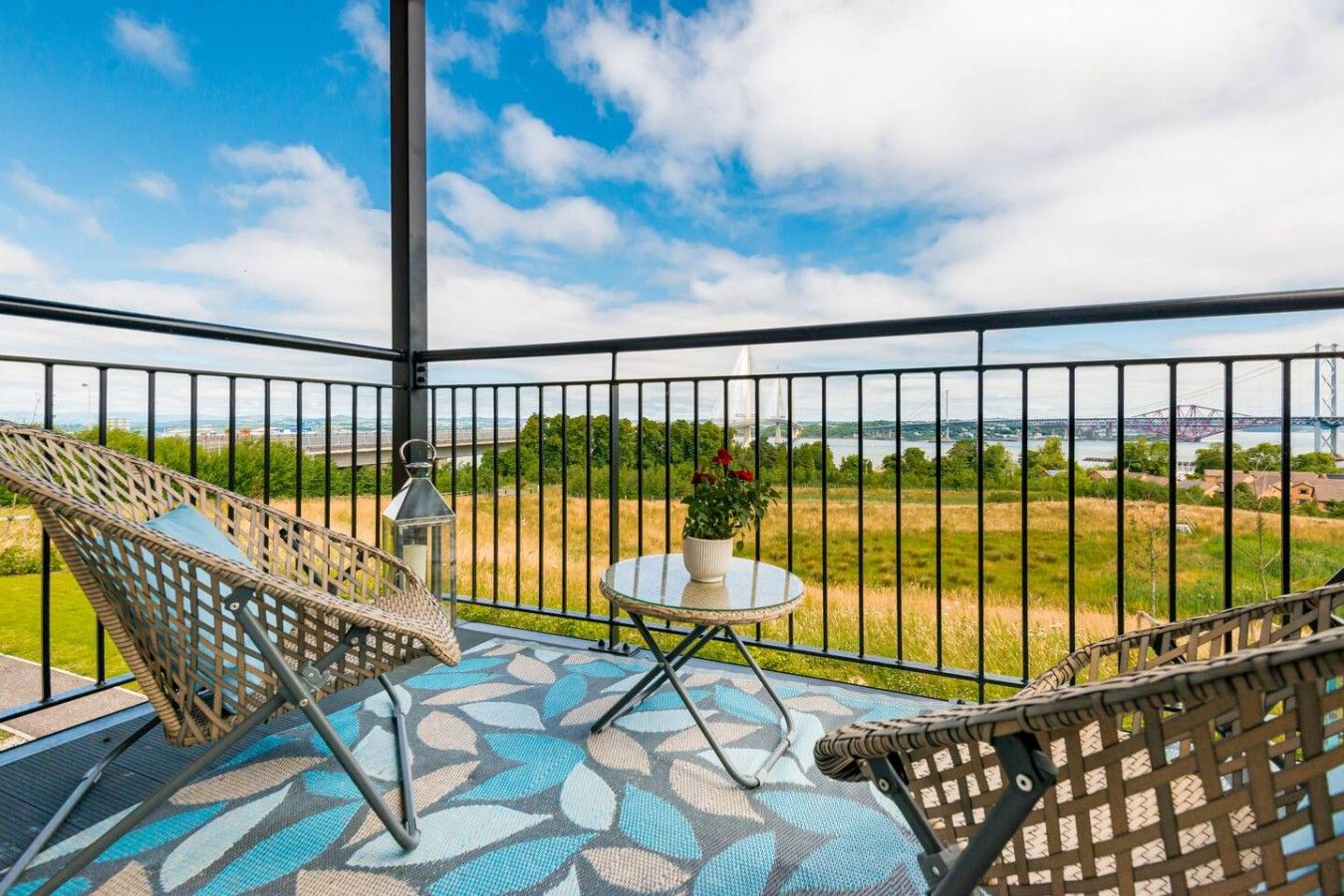 Ein Balkon mit zwei Korbstühlen, einem kleinen Tisch mit einem Blumentopf und Blick auf eine Brücke und Grünflächen.