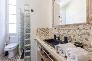 Ein Badezimmer mit Duschkabine aus Glas, Toilette, Waschbecken mit Rückwand aus Steinmosaik und Spiegel mit aufwendigem Rahmen.