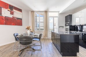 A modern kitchen with a central island, a dining area with a round table and chairs, and a large abstract painting on the wall. - Charming 2-Bedroom Apartment with Balcony in Forville A2B106 (Cannes)