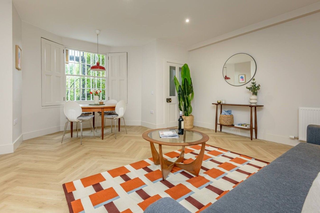 A modern living room with a dining area, a round coffee table, a large window with a view, and a mirror on the wall.