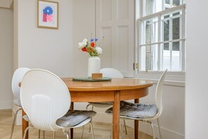 A dining area with a round wooden table, white chairs with cushioned seats, a vase with flowers, and a framed picture on the wall. - Central Stylish 1 Bed | Near Stockbridge (Edinburgh)