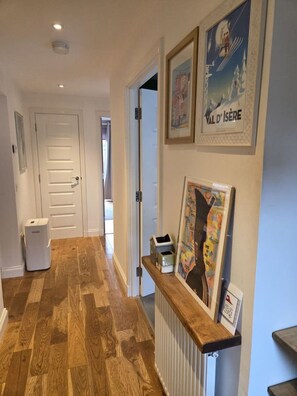 A hallway with wooden flooring, a white door, and a radiator. There are framed pictures on the wall and a small shelf with items. - Portobello Garden Flat | 2 bed Near Beach (Edinburgh)
