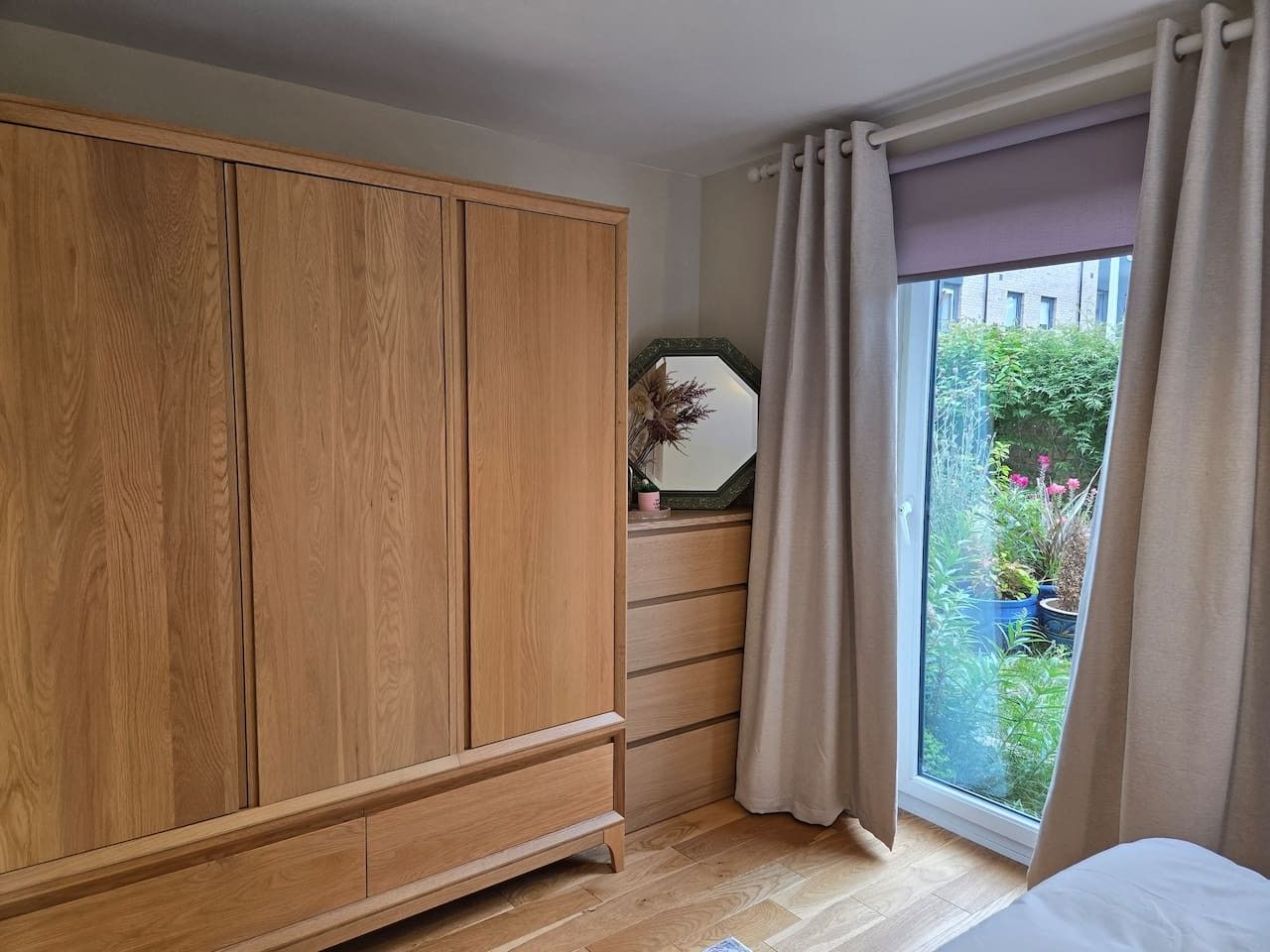 A bedroom with a wooden wardrobe, a bedside table with a mirror, and a window with curtains.