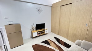 Ein modernes Wohnzimmer mit einem Flachbildfernseher auf einem Holzständer, einem kleinen weißen Schrank, einem braun-weißen Teppich und einem großen Kleiderschrank.