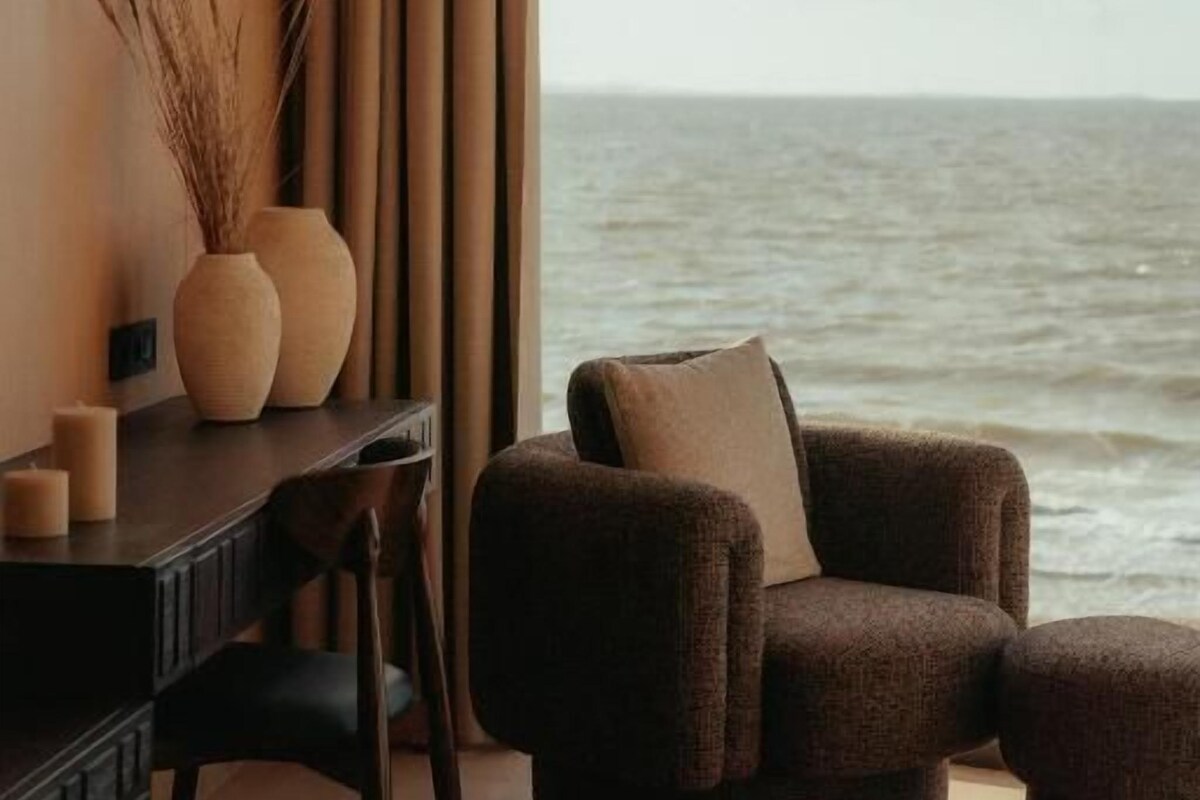 A room with a view of the sea, a round table, a sofa, and a desk with a vase and candles.