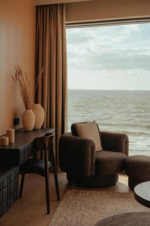 A room with a view of the sea, a round table, a sofa, and a desk with a vase and candles.