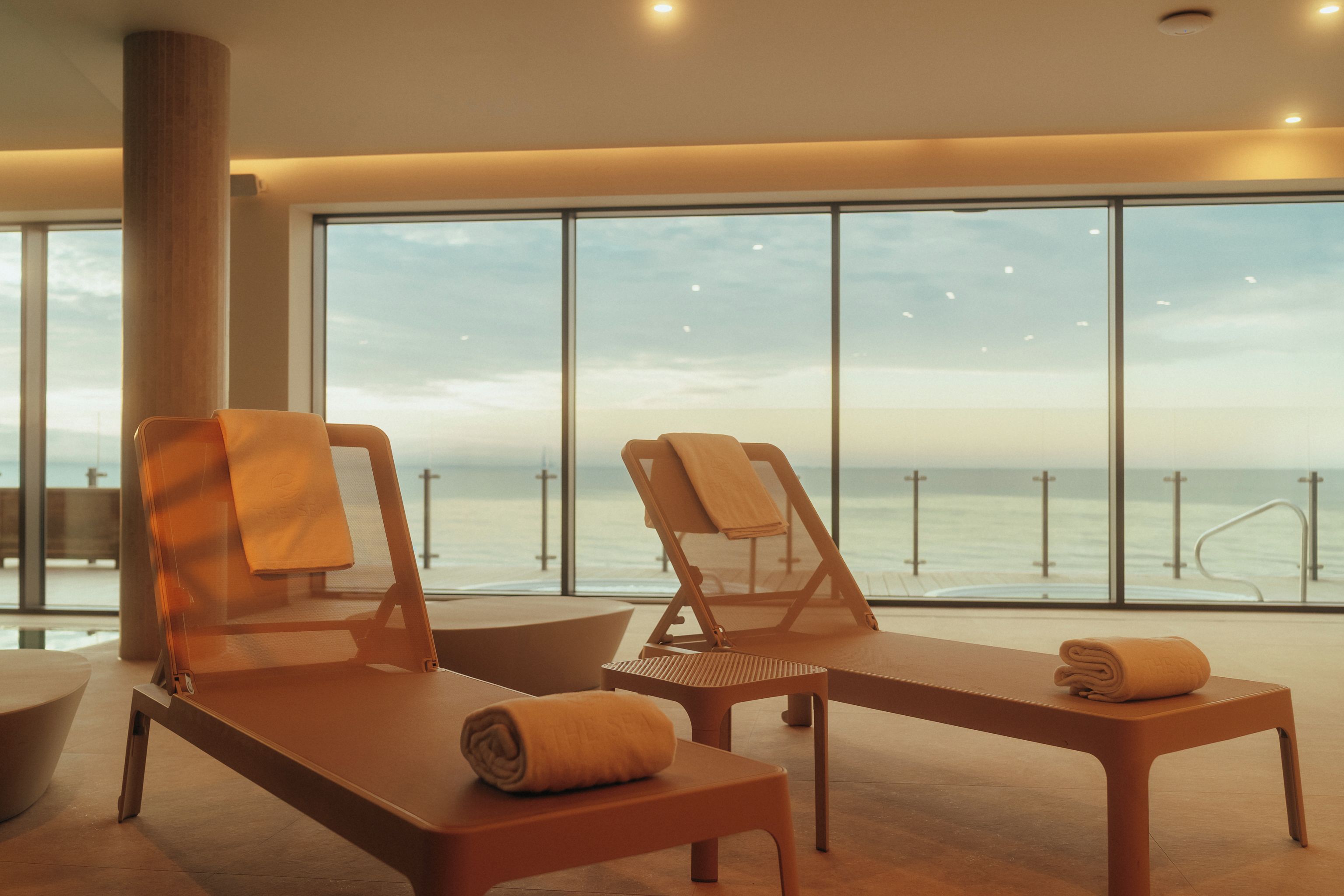 Een zonnige kamer met twee fauteuils en een bijzettafeltje, met uitzicht op een strand met een hekwerk.