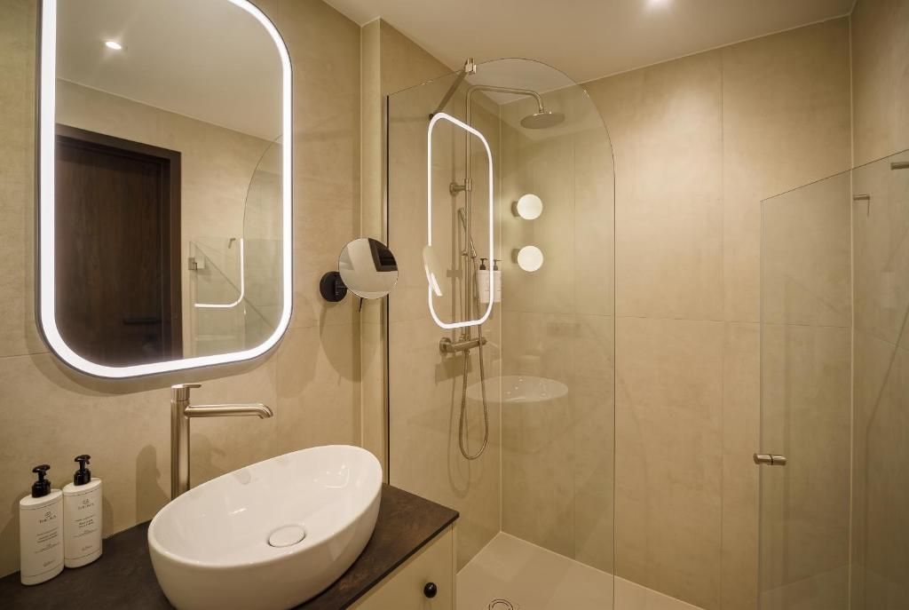 Un baño moderno con un lavabo ovalado grande, un espejo con iluminación integrada, una ducha con cerramiento de vidrio y una puerta que da a otra habitación.